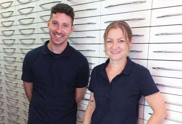 Zwei Mitglieder eines Herstellungsteams stehen lächelnd vor einer Wand mit Schubladen.