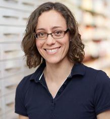 Eine Frau mit Brille und lockigem Haar lächelt freundlich in einer hellen Umgebung.