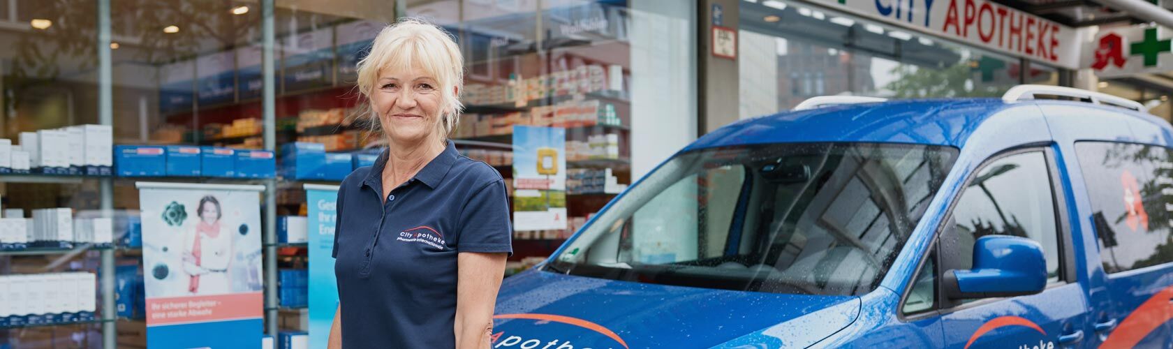 Eine Frau steht lächelnd vor einem blauen Lieferwagen, vor einer Apotheke, bereit für den Botendienst.