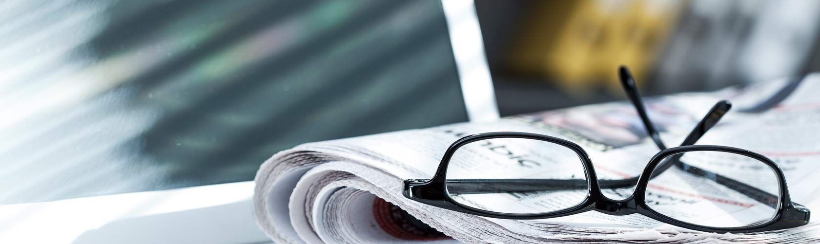 Eine schwarze Brille liegt auf einer gefalteten Zeitung, die im Sonnenlicht auf einem Tisch platziert ist.