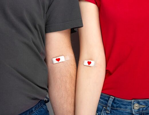 Zwei Menschen stehen nebeneinander, ihre Arme mit Pflastern und Herzsymbolen bedeckt, was auf Blutspende oder Haemophilie hinweist.
