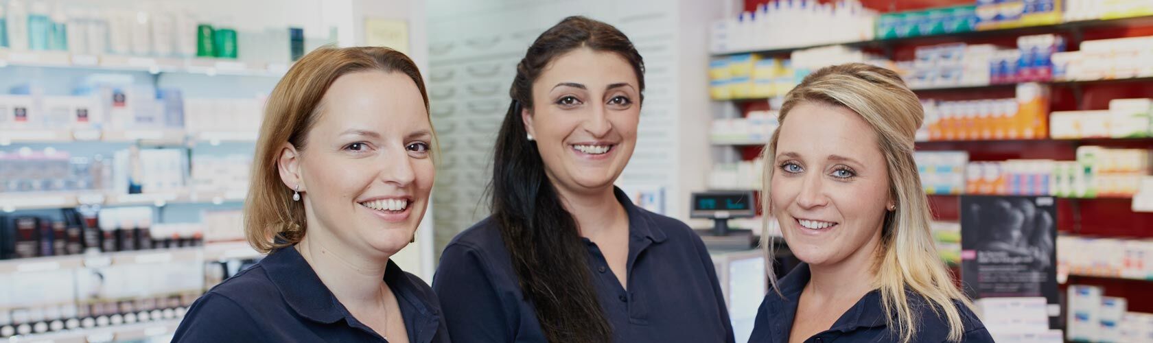 Drei Frauen in blauen Hemden lächeln in einer Apotheke, umgeben von Regalen voller Produkte, und fördern Aktionen.