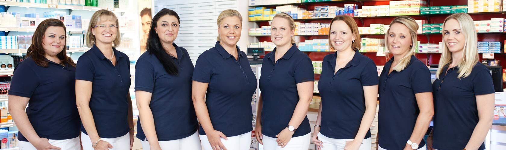 Acht Apothekenmitarbeiterinnen in blauen Poloshirts stehen lächelnd in einer Apotheke vor Regalen mit Medikamenten.